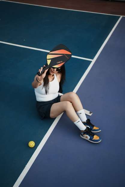 A player seated on a blue pickleball court, holding a paddle toward the camera